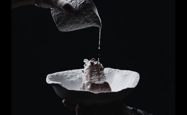 Dressage d’un dessert raffiné avec une sauce versée délicatement depuis un petit pichet, en noir et blanc, sur fond sombre.