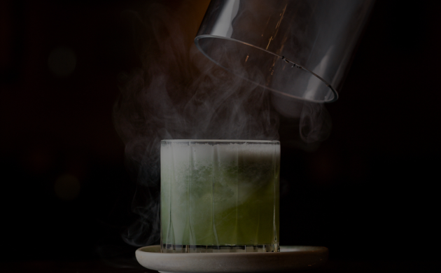 Cocktail vert fumant sous une cloche en verre sur fond noir.