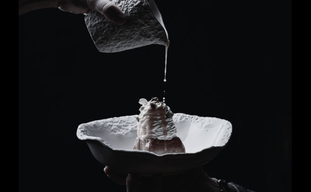 Dressage d’un dessert raffiné avec une sauce versée délicatement depuis un petit pichet, en noir et blanc, sur fond sombre.